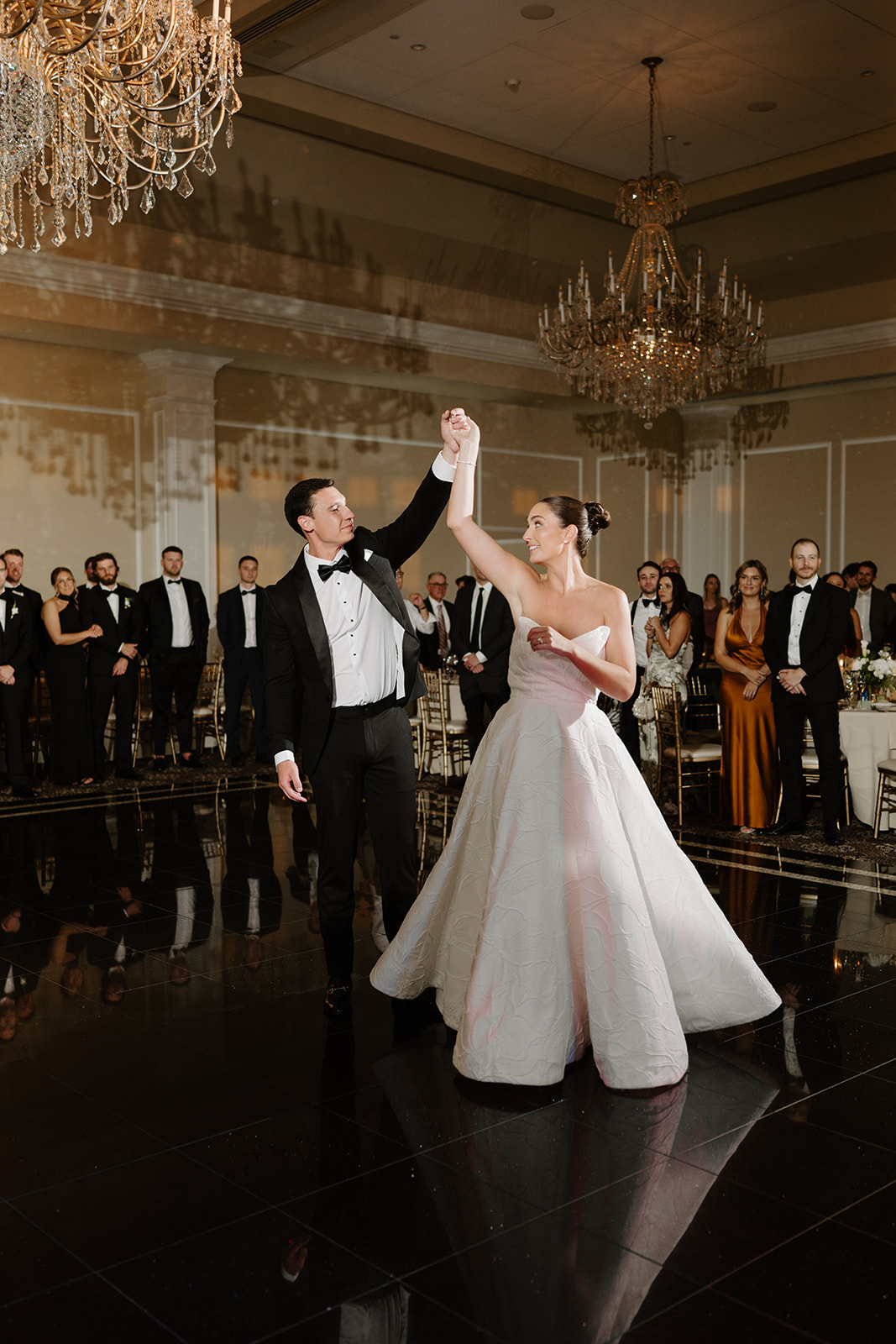 Groom spinning his smiling bride during their first dance on a reflective black ballroom floor.